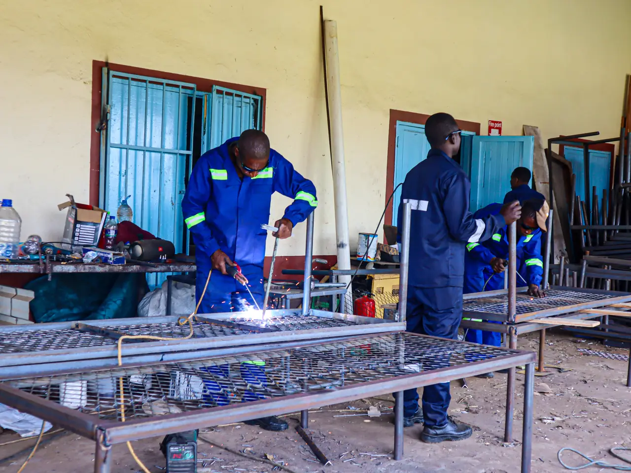 Learners in overalls welding steel bed frames in a school workshop
